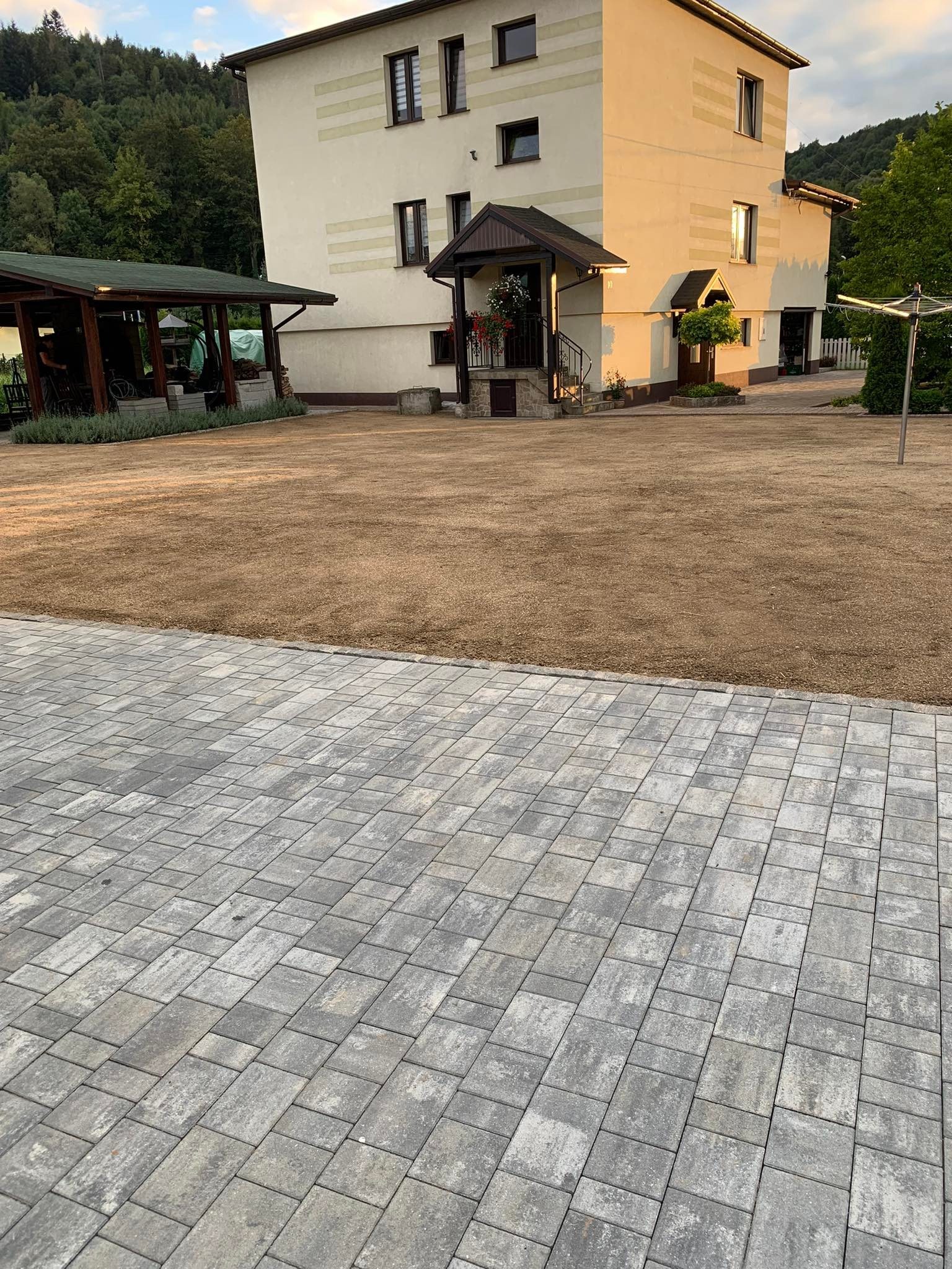 Nowo założony trawnik przed domem z szarą kostką brukową, widoczny budynek mieszkalny i altana w tle.