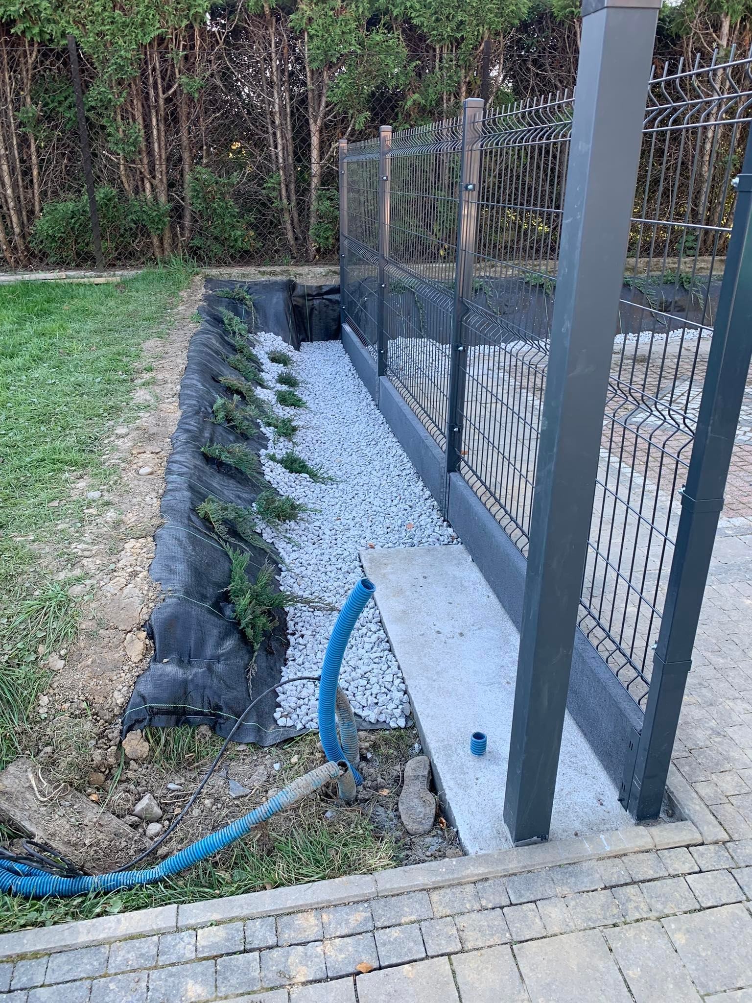 Przygotowany teren pod nasadzenia: wykop wyłożony czarną agrowłókniną, posadzone małe iglaki, wysypane kamieniami, widoczny fragment ogrodzenia panelowego.