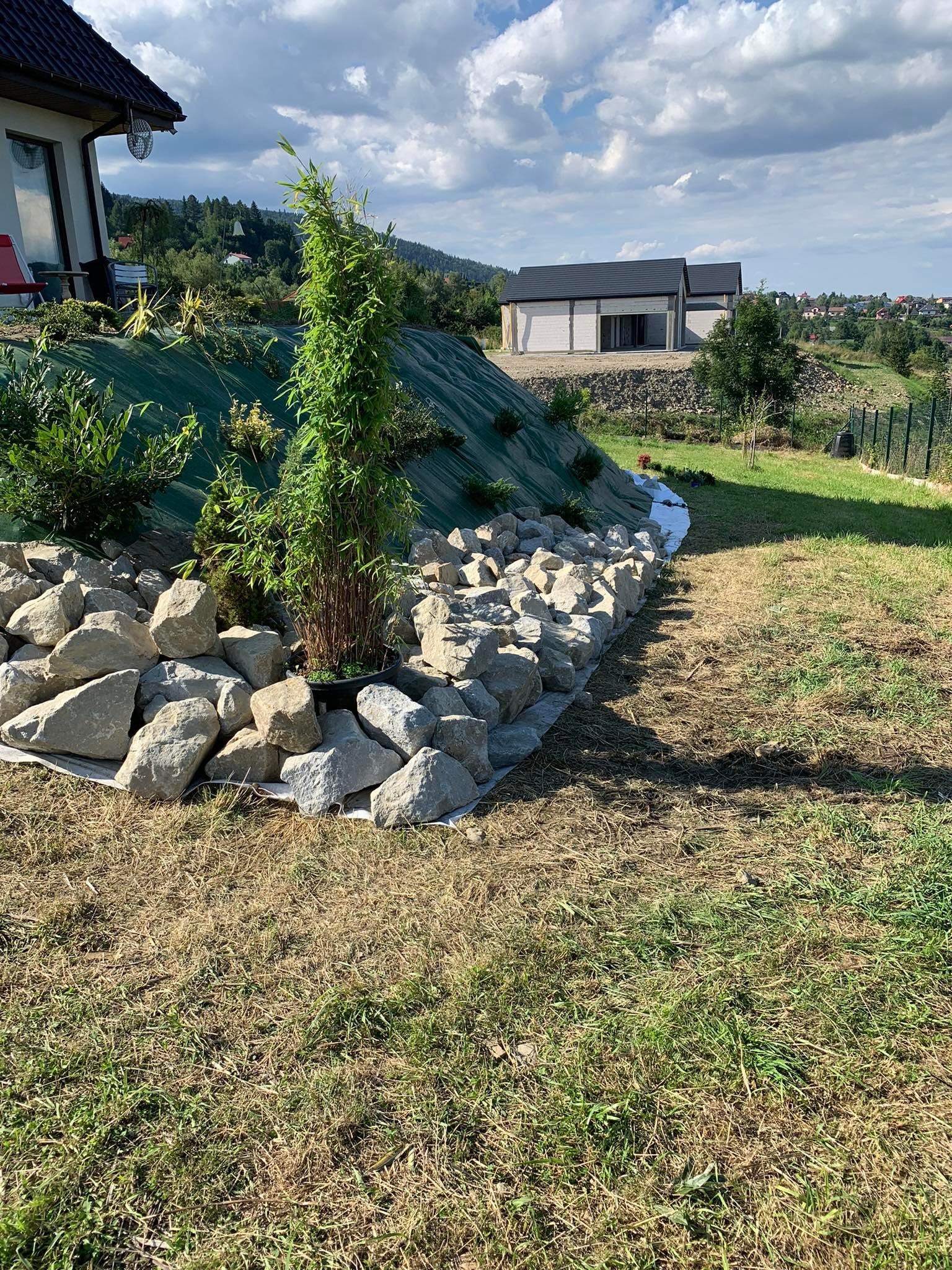 Aranżacja ogrodu z użyciem kamieni, bambusa w donicy i zielonej agrowłókniny na zboczu, widok na domy w oddali.