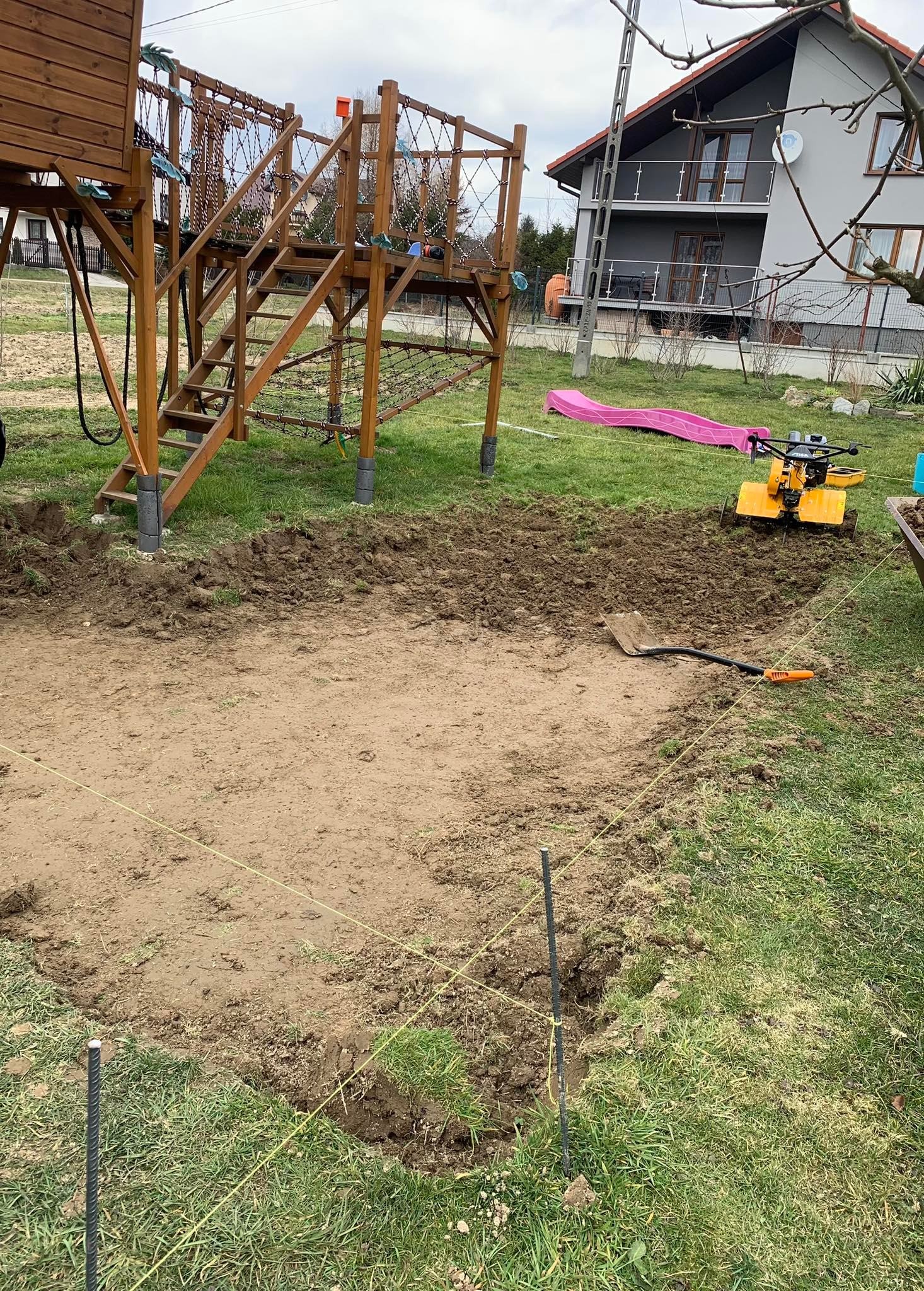 Przygotowany teren pod trawnik z wytyczonym obrysem, widoczny glebogryzarka, szpadel i paliki z linką, w tle plac zabaw i dom.