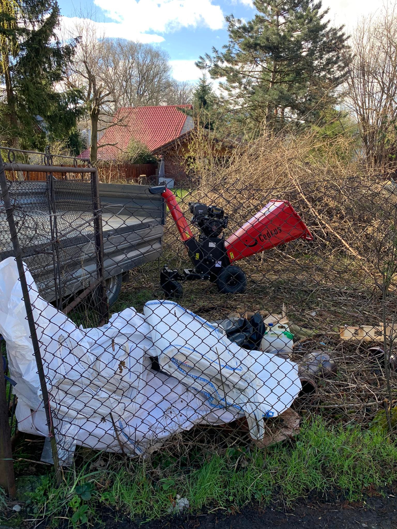 Czerwona rębarka Cedrus za metalową siatką, obok przyczepa i worki z białym materiałem, w tle budynek z czerwonym dachem i drzewa bez liści.
