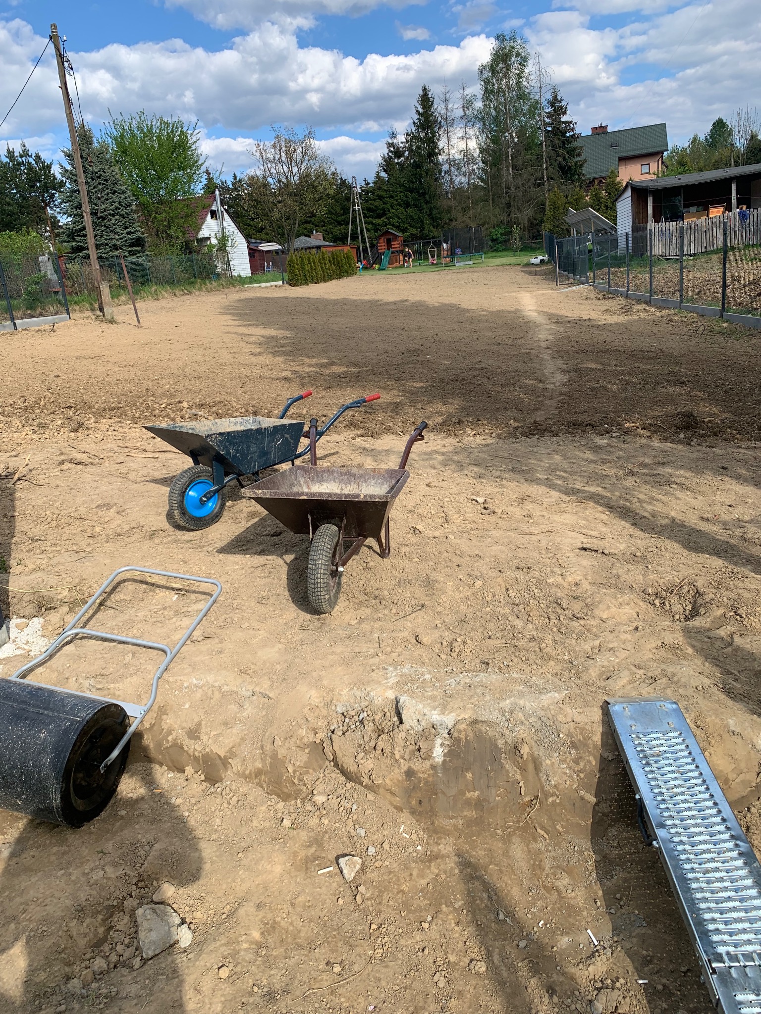 Przygotowany teren pod trawnik w Żywcu, z widocznymi dwoma taczkami, walcem i kratką drenażową, gotowy do dalszych prac.