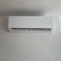 Nowoczesny biały klimatyzator marki Gree zamontowany na białej ścianie, widoczny fragment okna i budynek w tle.