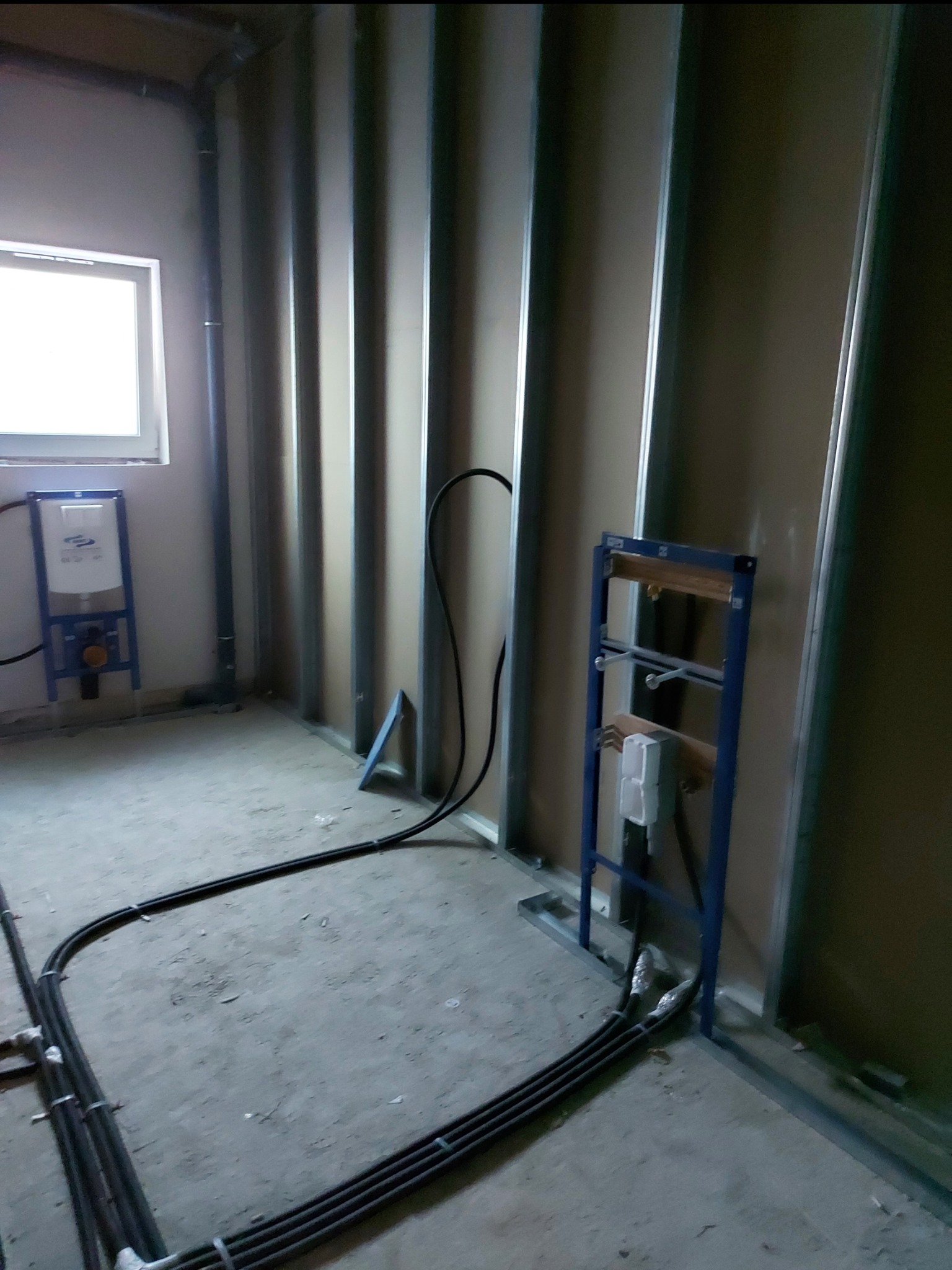 Wnętrze łazienki w trakcie prac: widoczne profile ścianek działowych, rury instalacyjne ułożone na podłodze i stelaż podtynkowy WC z zaworem, okno z dostępem światła dziennego.