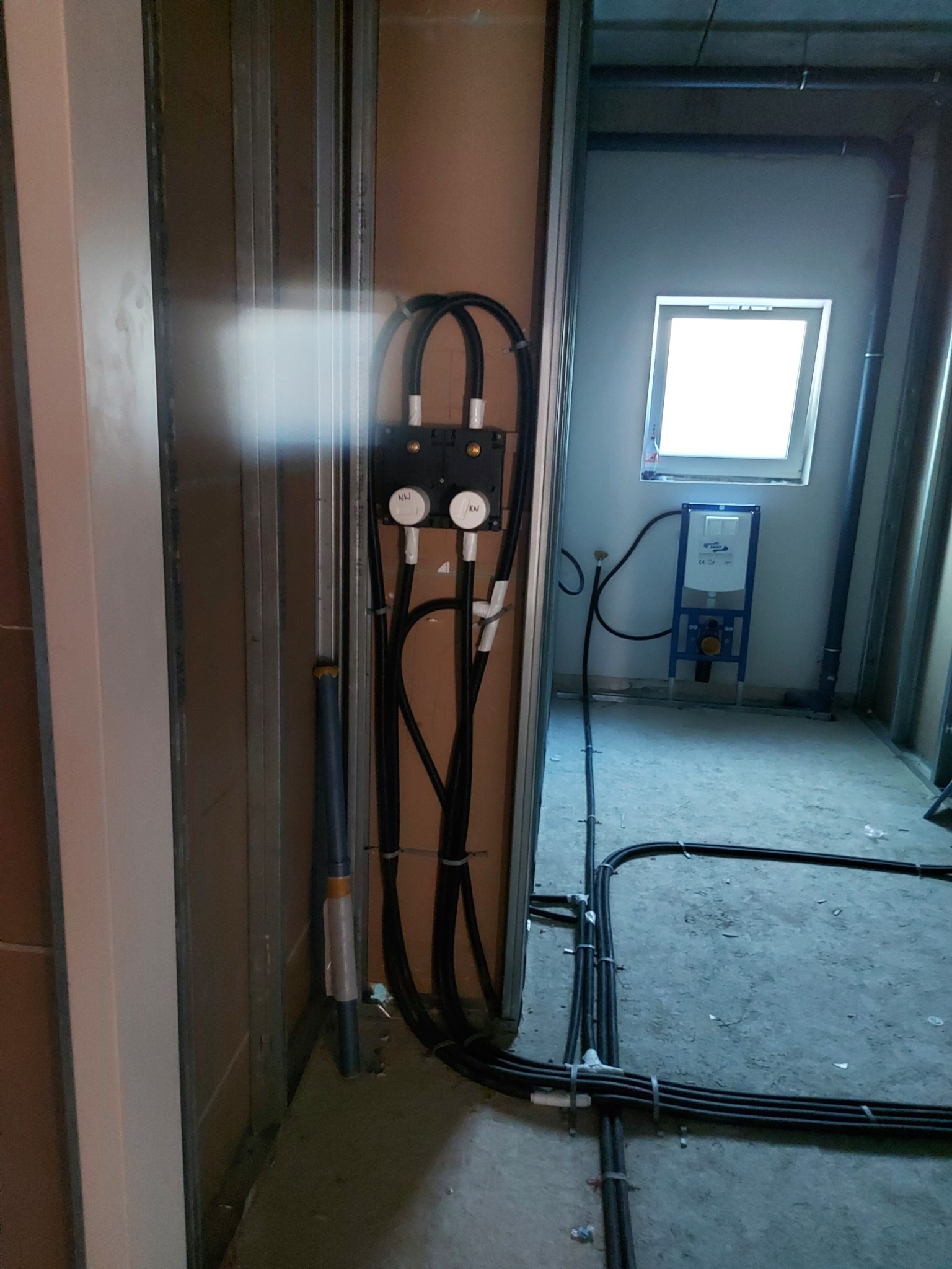 Wnętrze łazienki w trakcie prac instalacyjnych; widoczne przewody elektryczne, rury kanalizacyjne, stelaż podtynkowy WC przy oknie oraz surowa konstrukcja ścian z profili.