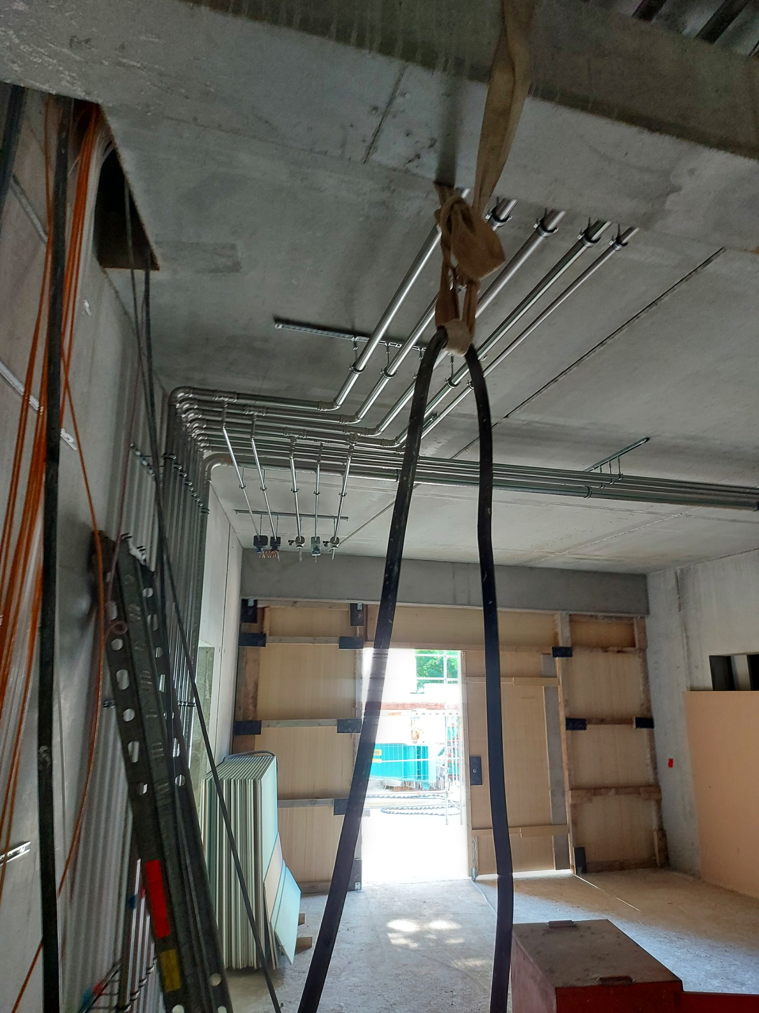 Surowy betonowy sufit z zamontowanymi metalowymi rurami instalacji, z wiszącymi kablami w osłonie, widok na otwarte drzwi i rusztowanie w tle.