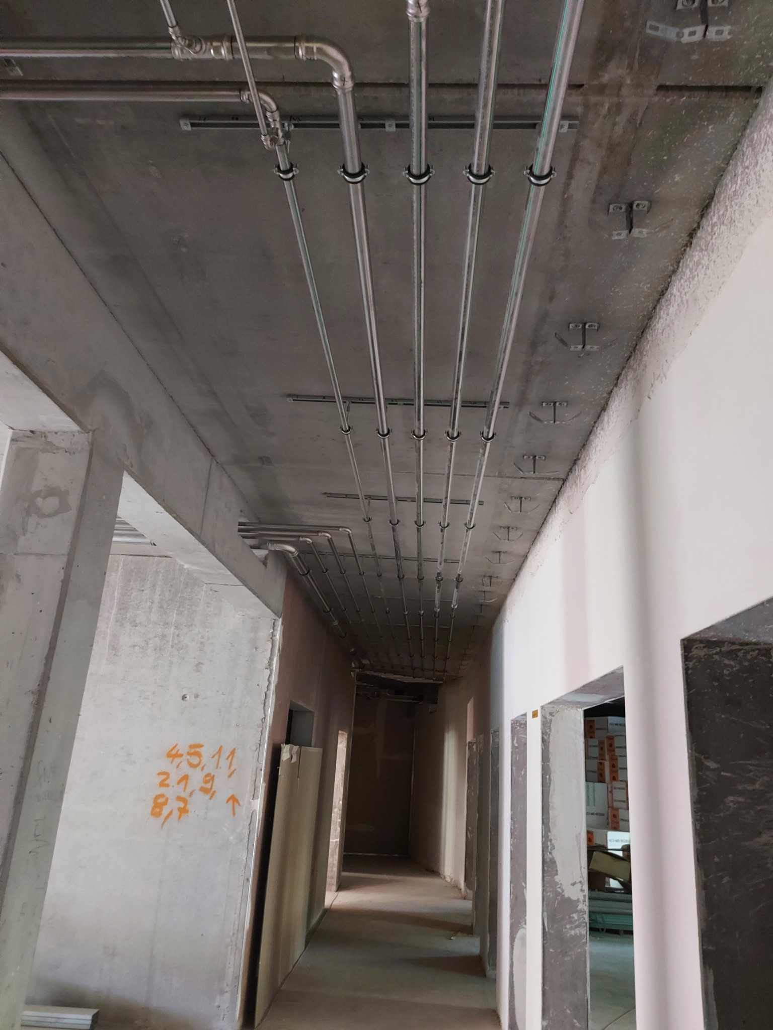 Montaż instalacji rurowej pod betonowym sufitem w surowym wnętrzu, widoczne metalowe rury mocowane klamrami, pomarańczowe napisy na ścianie w tle.
