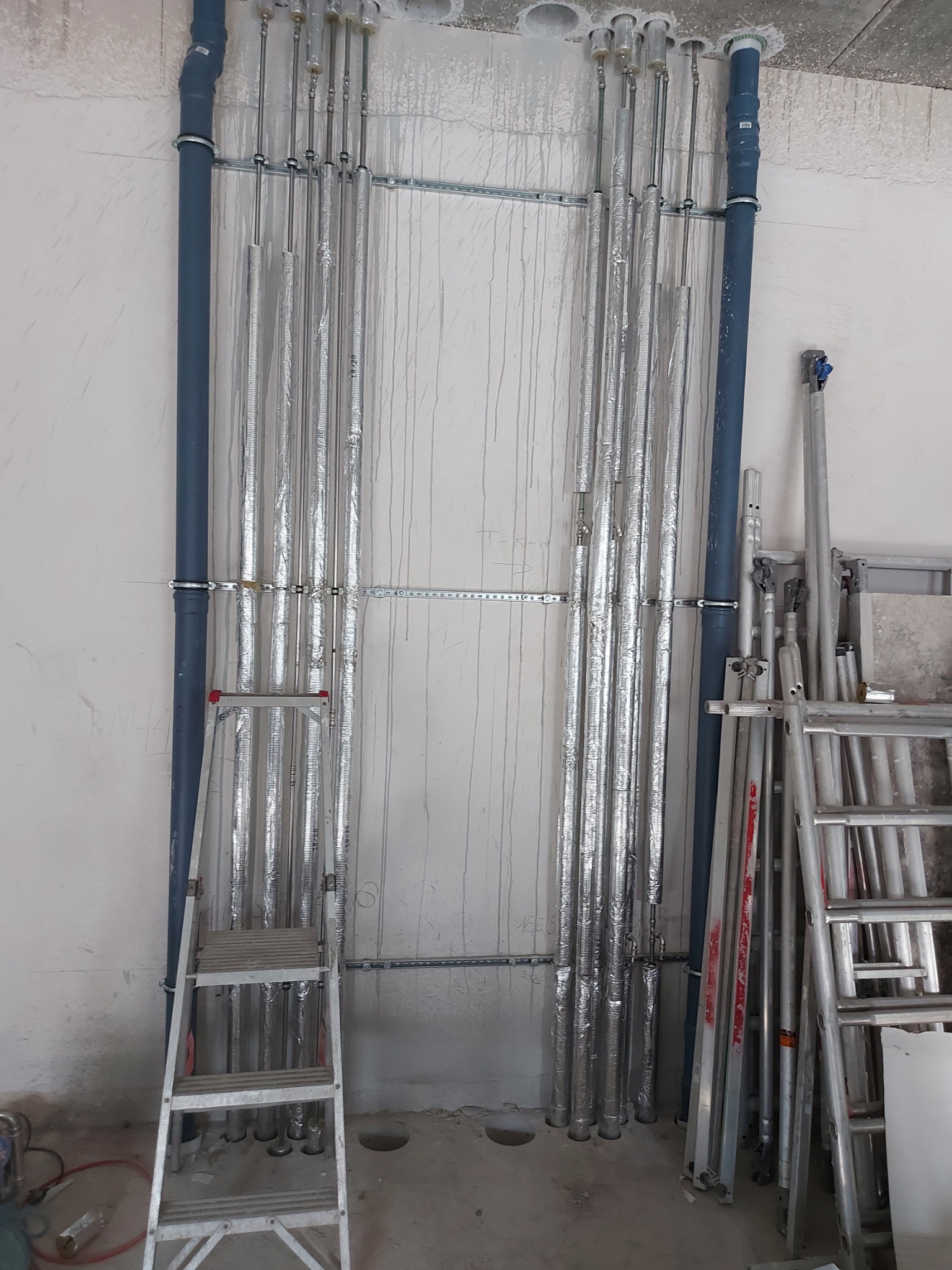Instalacja rur w folii izolacyjnej, mocowanych do ściany z widocznymi śladami po wierceniu, z drabinką aluminiową opartą o rury.