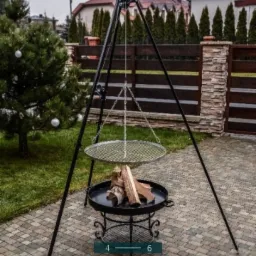 Kociołek myśliwski na trójnogu z rusztem, z ułożonym drewnem, na tle kostki brukowej, ogrodzenia i domu.