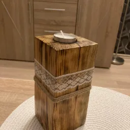 Drewniany świecznik w kształcie sześcianu, ozdobiony koronką i sznurkiem, z umieszczoną na górze tealight, na tle mebli w kolorze drewna.