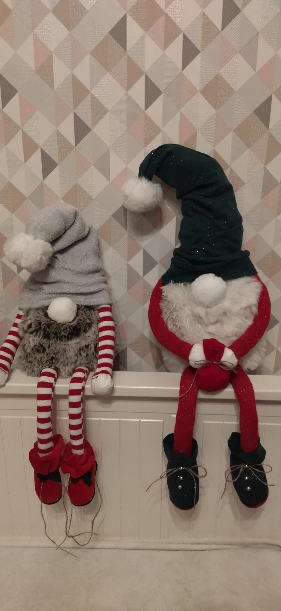 Dwa dekoracyjne gnomy siedzące na białej półce na tle ściany z geometrycznym wzorem. Jeden gnom ma szary kapelusz i czerwono-białe paski na nogach, drugi ma zielony kapelusz i czerwone nogi.