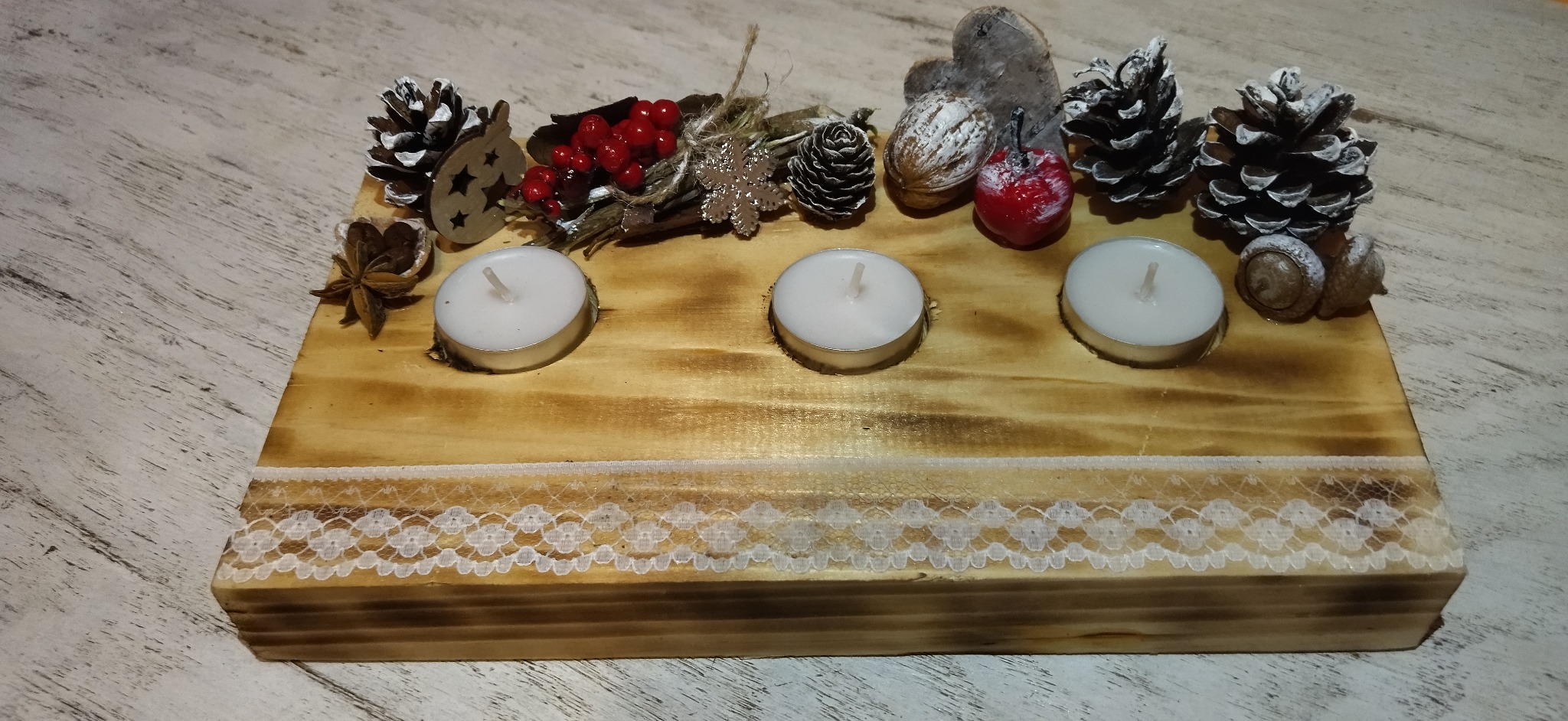 Drewniany świecznik z trzema tealightami, ozdobiony szyszkami, jarzębiną i koronką, na tle imitującym bielone drewno.