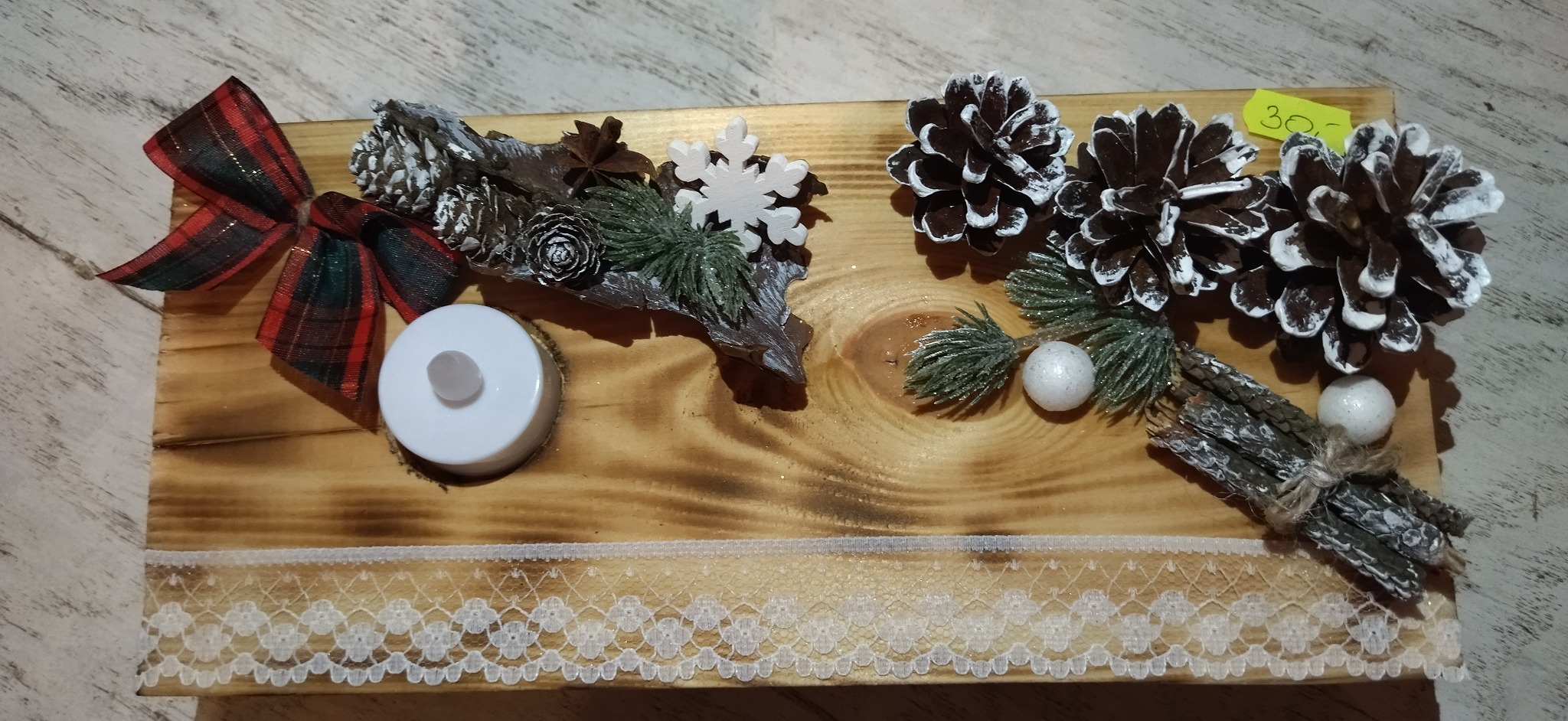 Dekoracja świąteczna DIY na drewnianej desce z koronkowym wykończeniem, obejmująca szyszki z białymi końcówkami, gałązki świerkowe, kokardę w kratę, drewniane ozdoby i świecę LED.