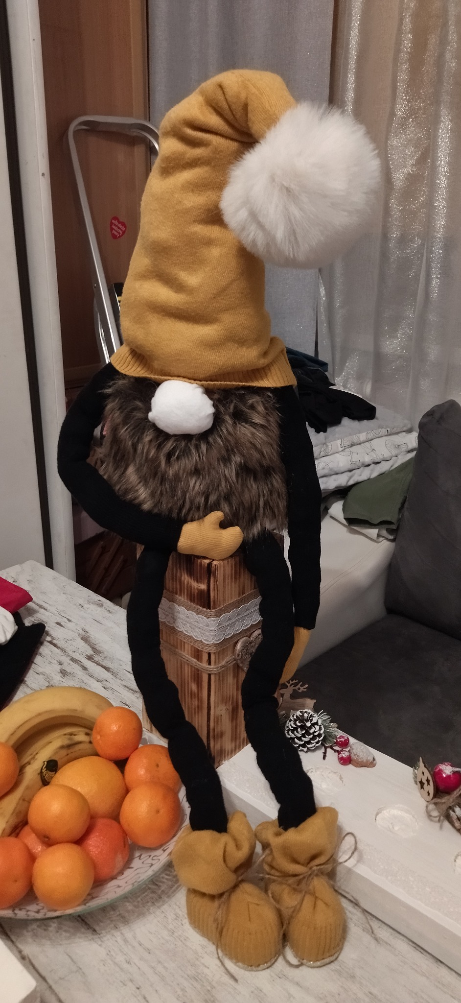 Dekoracyjny skrzat z żółtą czapką z pomponem, futrzaną brodą i czarnymi nogami, siedzący na drewnianej skrzynce z koronką, otoczony owocami i świątecznymi ozdobami.