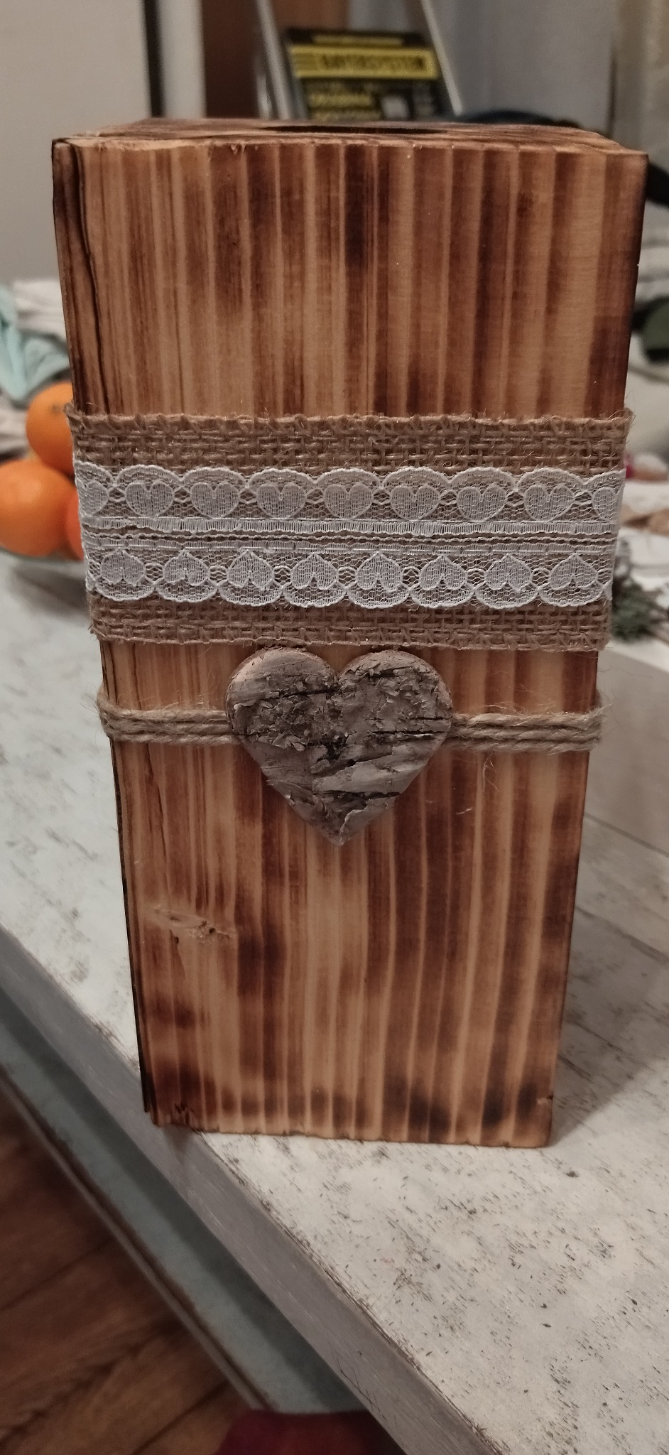 Ręcznie wykonana, drewniana osłonka na doniczkę, zdobiona jutowym sznurkiem, koronką z motywem serca i drewnianym serduszkiem z kory brzozy, na tle widoczne pomarańcze.