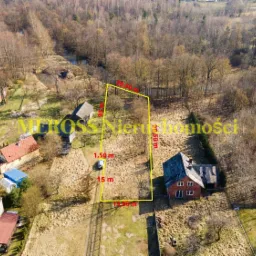 Działka budowlana z naniesionymi wymiarami i granicami, widok z drona, otoczona drzewami i zabudową jednorodzinną.