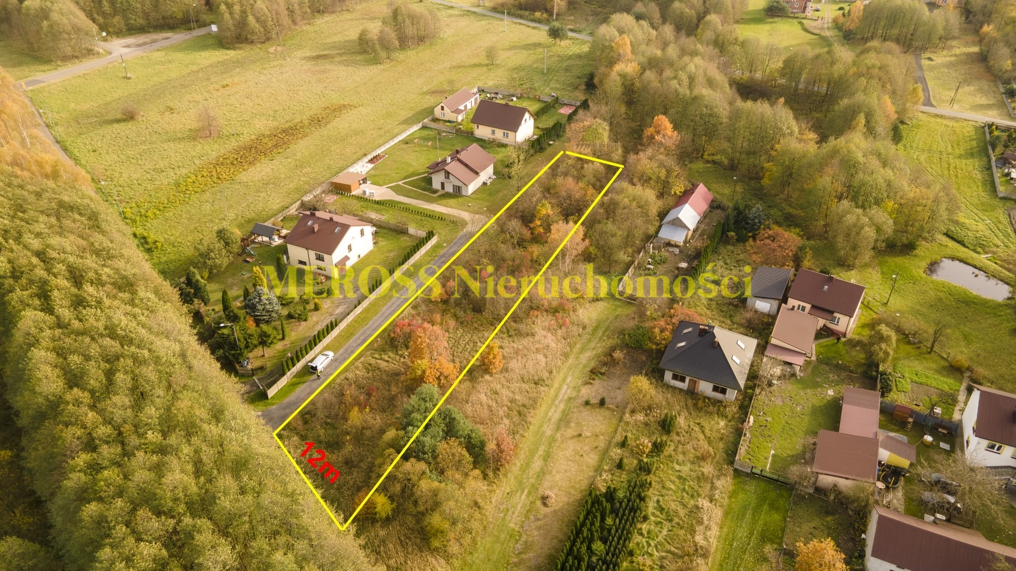 Zdjęcie z lotu ptaka przedstawiające działkę budowlaną otoczoną żółtą linią, z widocznymi domami jednorodzinnymi i drzewami w jesiennych kolorach. Na zdjęciu widoczne jest logo firmy nieruchomości.