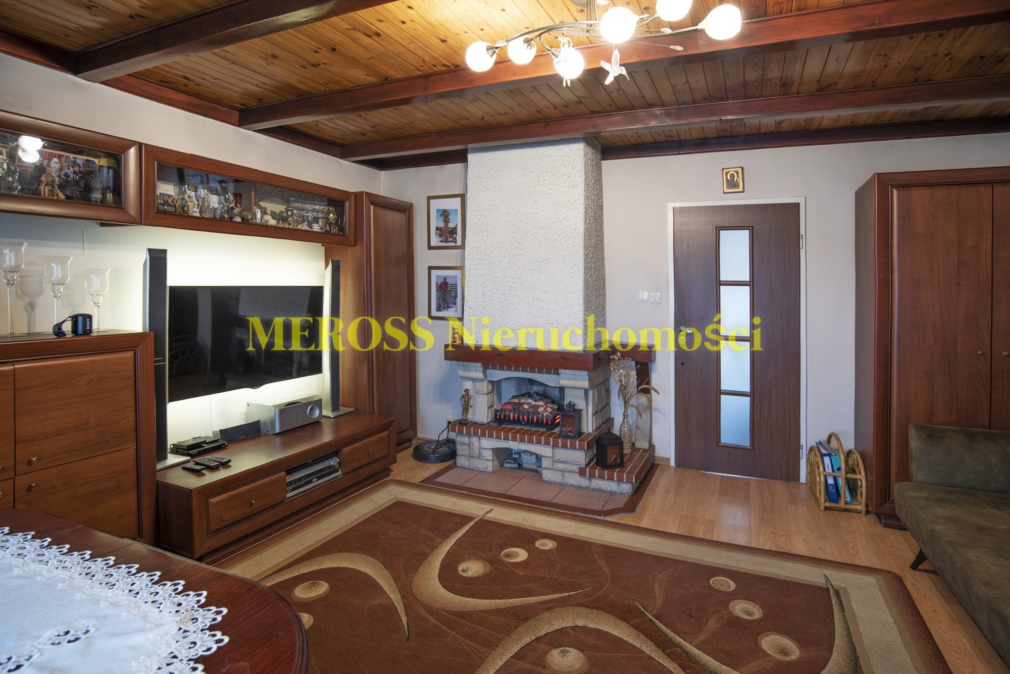 Jasne wnętrze salonu z drewnianym sufitem, kominkiem z paleniskiem, brązowymi meblami i wzorzystym dywanem.