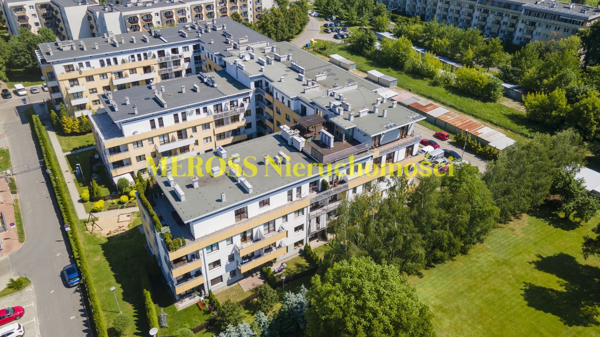 Widok z lotu ptaka na kompleks nowoczesnych budynków mieszkalnych z balkonami i zielonym otoczeniem, w tle widoczne parkingi i drzewa.