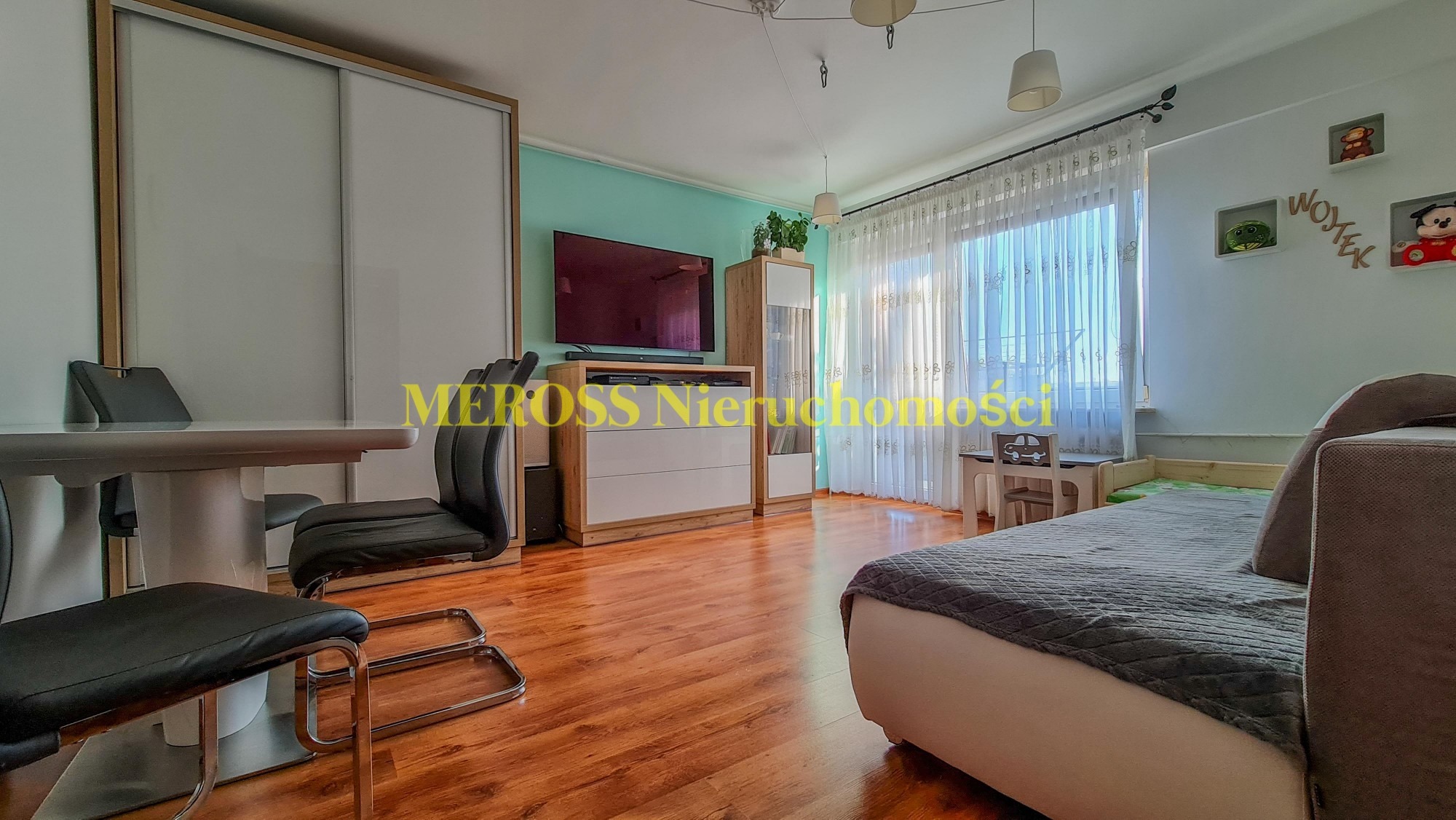 Jasne wnętrze pokoju z drewnianą podłogą, biurkiem dla dziecka pod oknem i rozkładaną sofą; widoczne również stół z krzesłami oraz szafa z przesuwnymi drzwiami.