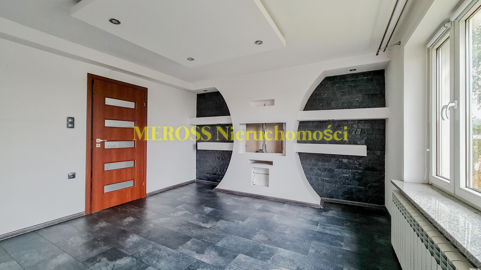 Puste pomieszczenie z ciemną podłogą, białymi ścianami i dekoracyjną ścianą z czarną okładziną imitującą kamień, wnęka na kominek bez wkładu, brązowe drzwi z przeszkleniami i okno z marmurowym...