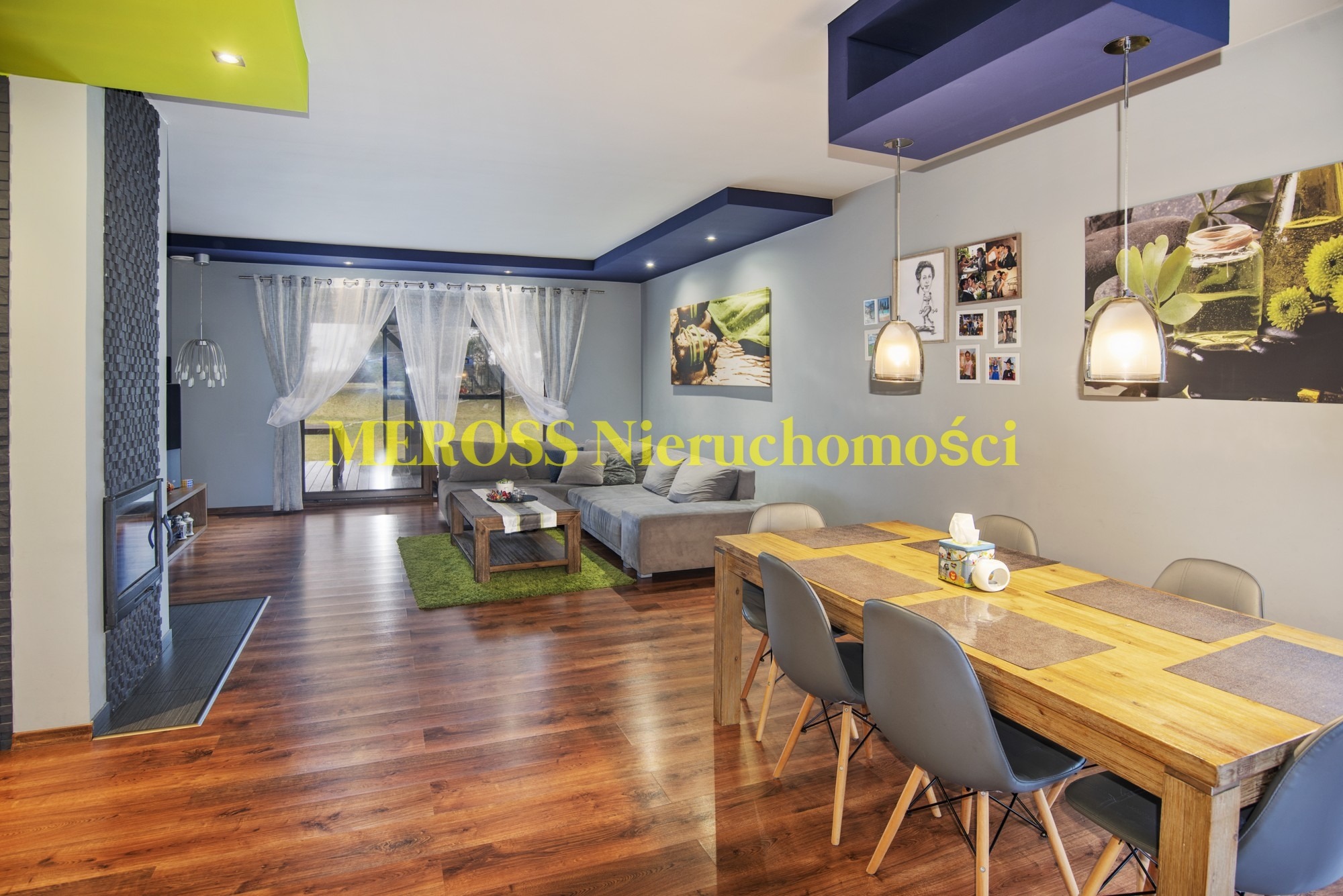 Jasne, przestronne wnętrze salonu z drewnianą podłogą, szarą sofą narożną, drewnianym stołem jadalnym z szarymi krzesłami, kominkiem z kamienną okładziną i zielonym akcentem kolorystycznym...