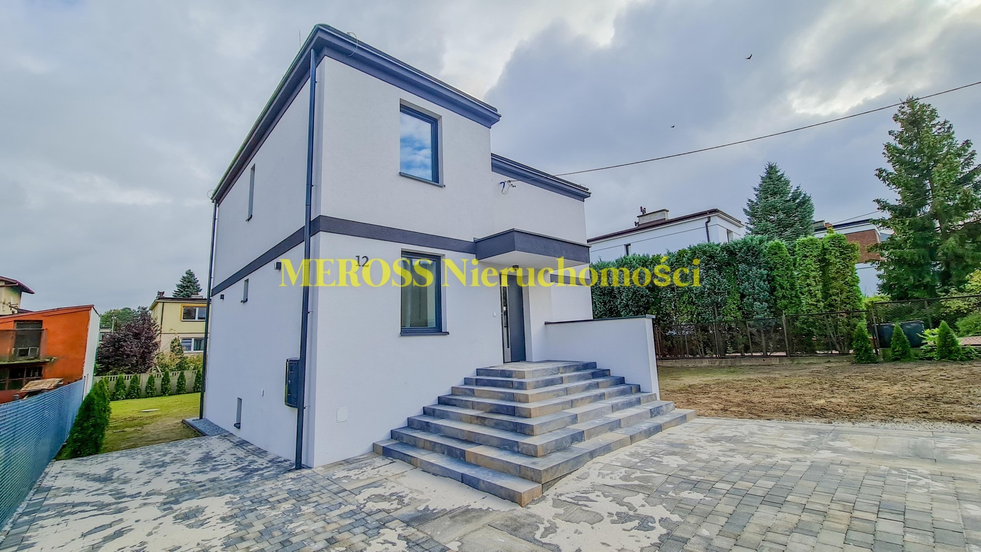 Nowoczesny, dwukondygnacyjny dom z białymi ścianami, szarymi akcentami i szerokimi schodami wejściowymi z kostki brukowej.