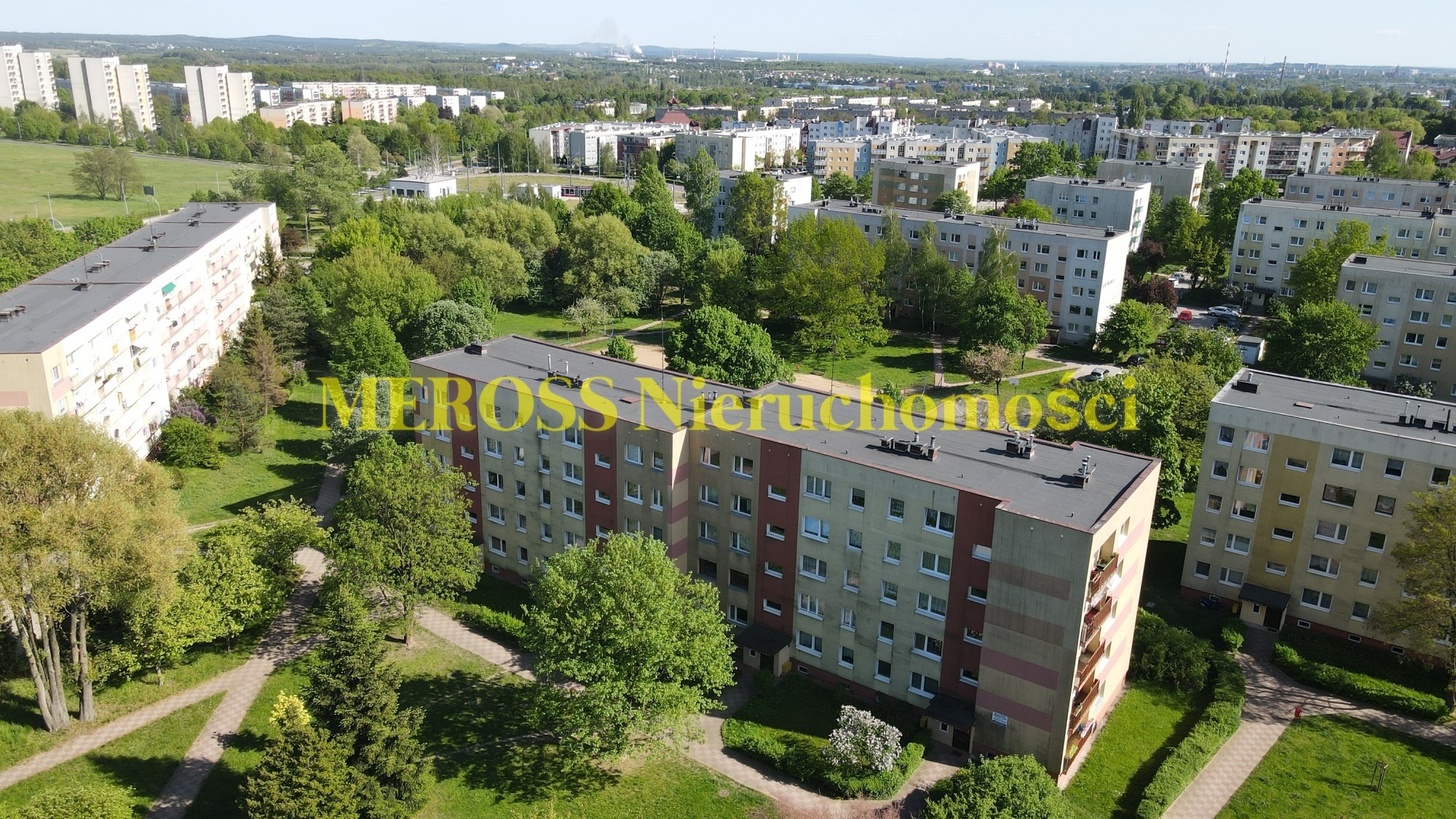 Widok z góry na osiedle mieszkaniowe z blokami z lat 80-tych, otoczone zielenią, w tle zabudowa miejska i komin