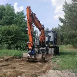 Pomarańczowa koparka kołowa Hitachi z łyżką w trakcie prac ziemnych na terenie zarośniętym trawą, w tle drzewa liściaste i iglaste, operator w kabinie.