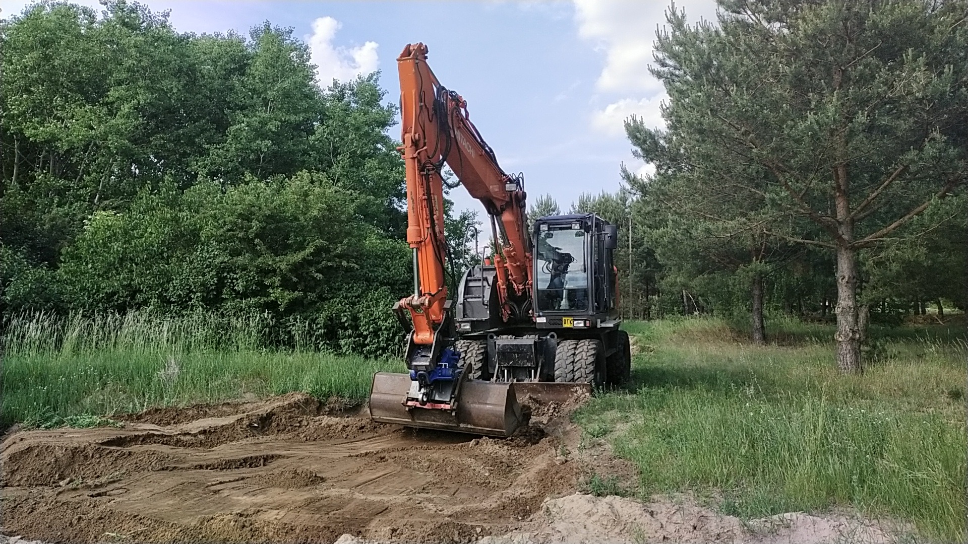 Pomarańczowa koparka kołowa Hitachi z łyżką w trakcie prac ziemnych na terenie zarośniętym trawą, w tle drzewa liściaste i iglaste, operator w kabinie.