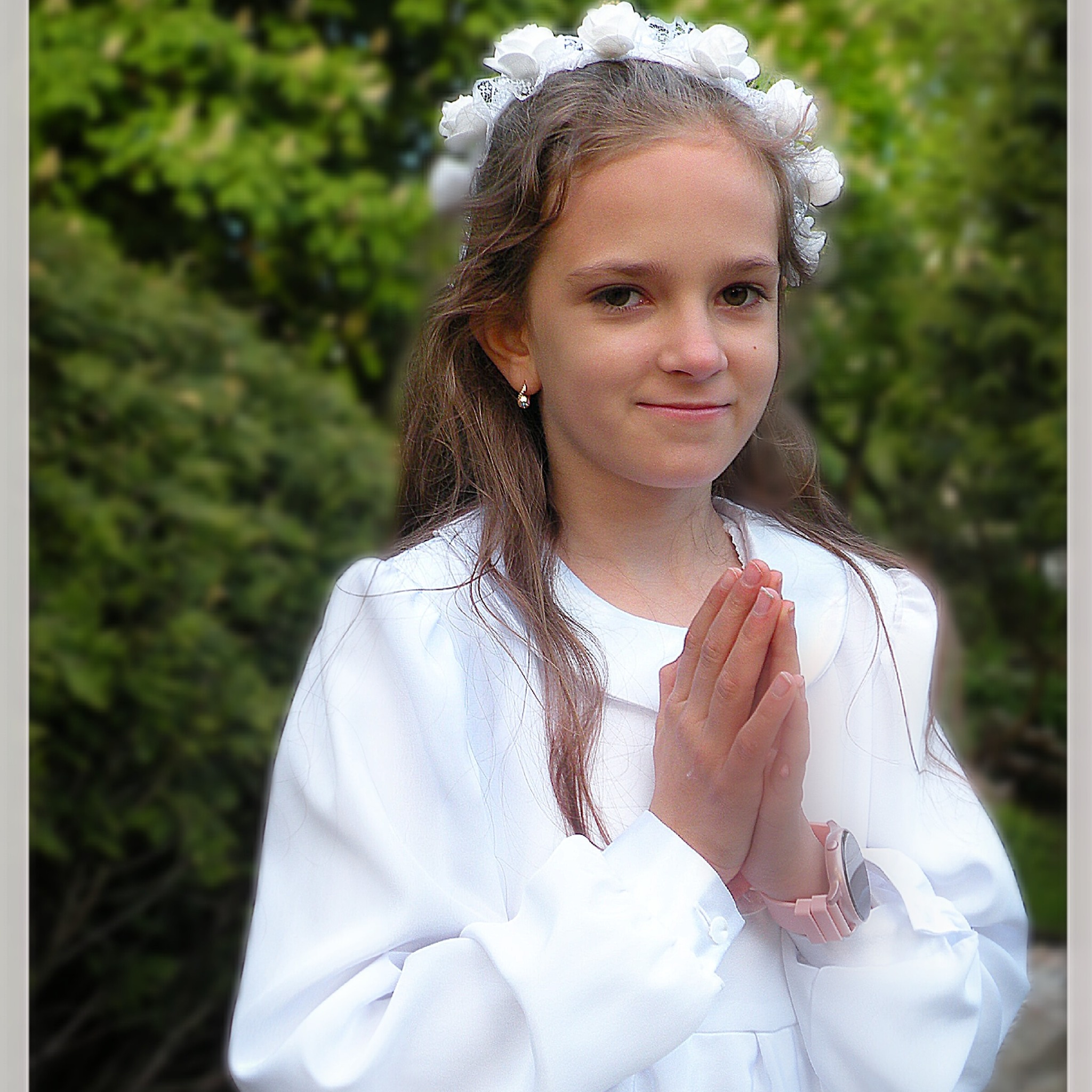 Dziewczynka w białej albie i wianku z białych kwiatów, z rękami złożonymi do modlitwy, na tle zieleni.