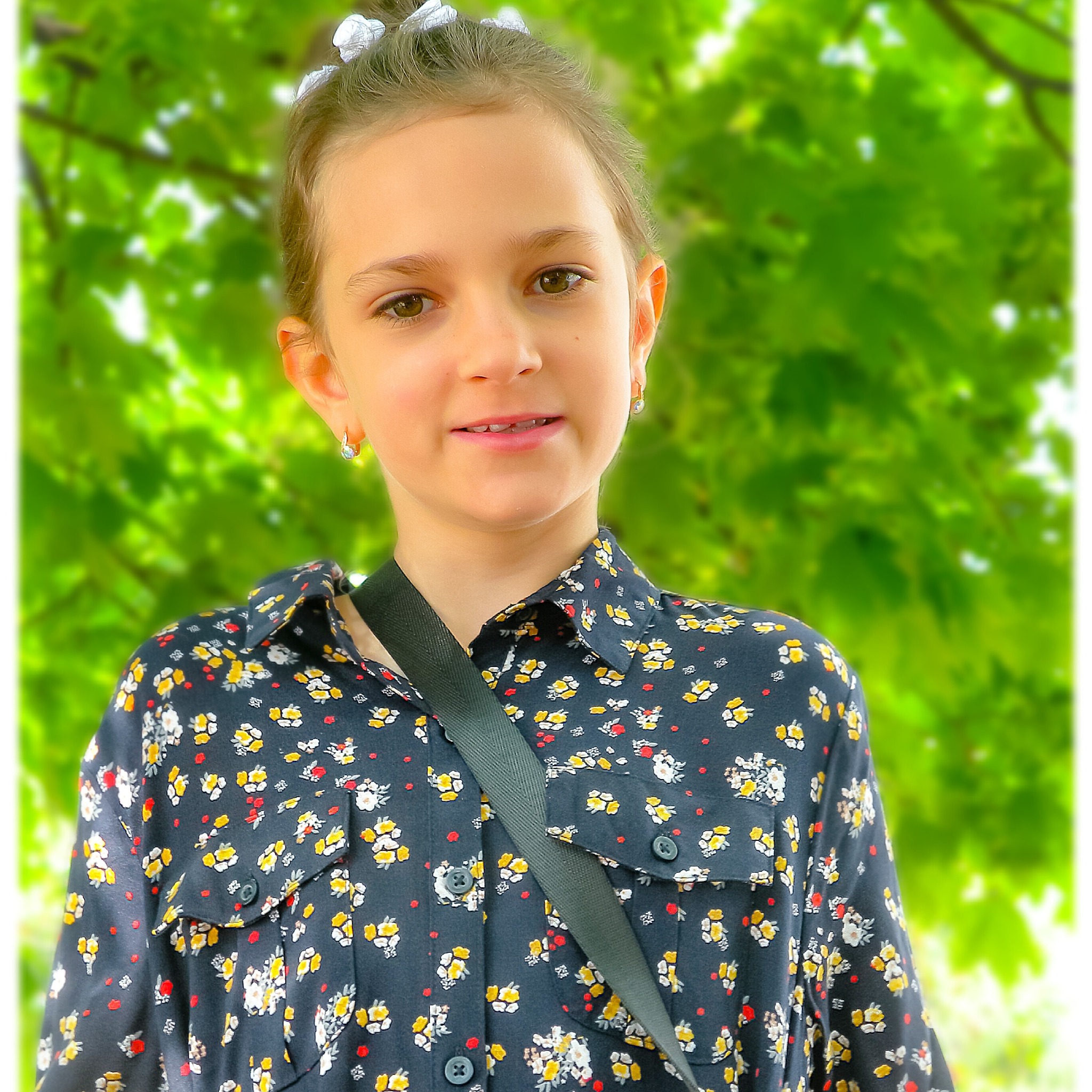 Portret uśmiechniętej dziewczynki w kwiecistej koszuli z paskiem na ramieniu, na rozmytym tle zielonych liści.