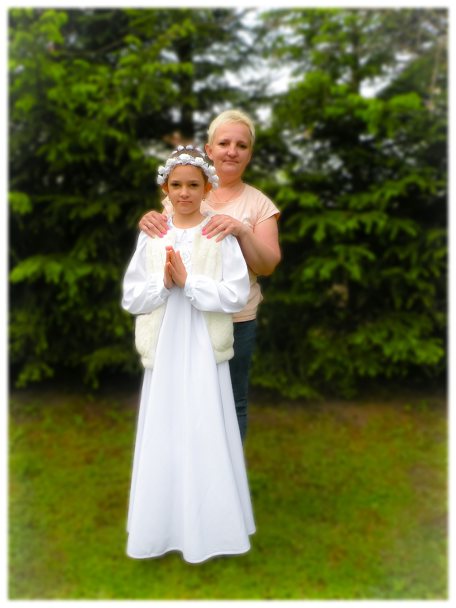 Dziewczynka w białej albie komunijnej z wiankiem na głowie, stojąca na tle zieleni, z rękami złożonymi do modlitwy, wspierana przez kobietę z tyłu.