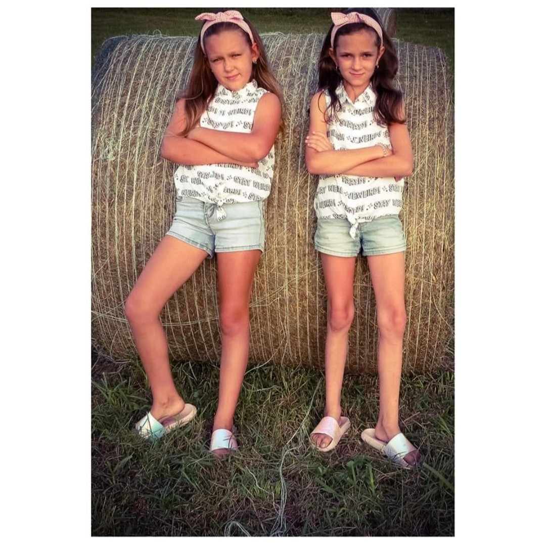 Dwie dziewczynki w identycznych strojach, stojące na trawie przed stogiem siana. Ubrania w biało-czarne wzory, krótkie spodenki i opaski z kokardkami. Uśmiechnięte, z założonymi rękoma.