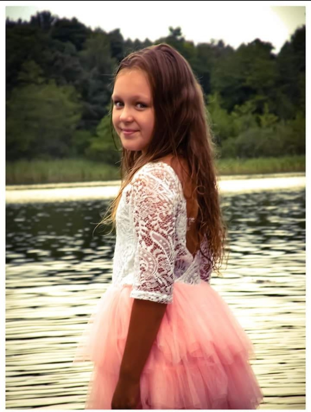 Uśmiechnięta dziewczynka w koronkowej bluzce i różowej tiulowej spódniczce, pozuje na tle jeziora i zieleni. Delikatne światło podkreśla naturalność scenerii.