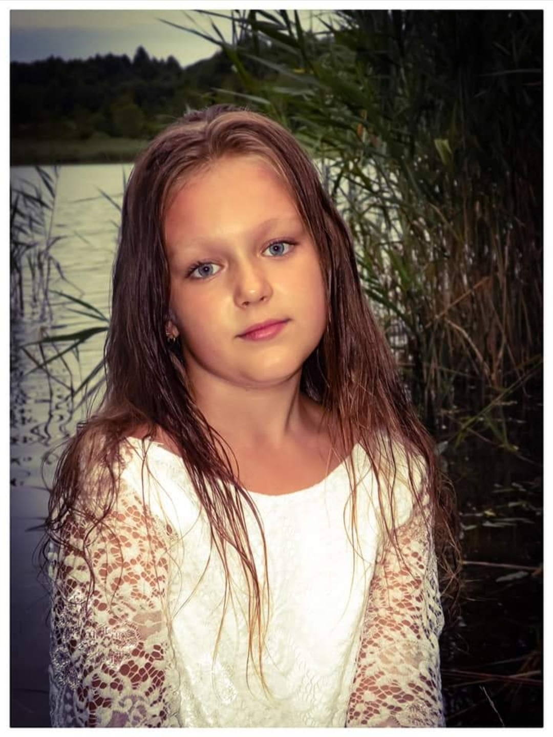 Portret młodej dziewczyny w białej koronkowej sukience na tle jeziora i trzcin, z mokrymi włosami opadającymi na ramiona.