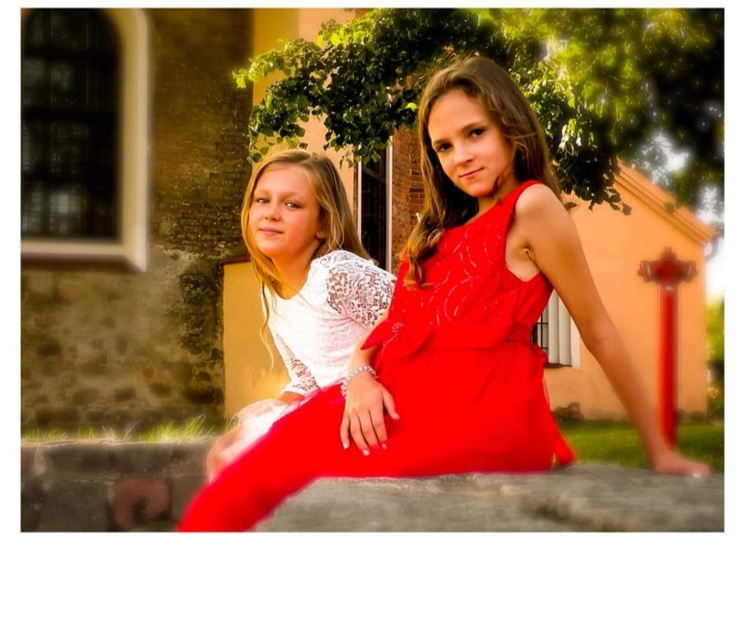 Dwie dziewczynki pozują na tle starego budynku i zieleni. Jedna w białej koronkowej bluzce, druga w czerwonej sukience. Delikatne światło słoneczne podkreśla ich naturalność.