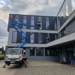 Pracownik w niebieskim podnośniku koszowym myje okna na wysokości w nowoczesnym biurowcu z logo EKOMA na dachu; biała ciężarówka z podpórkami stabilizującymi na kostce brukowej.