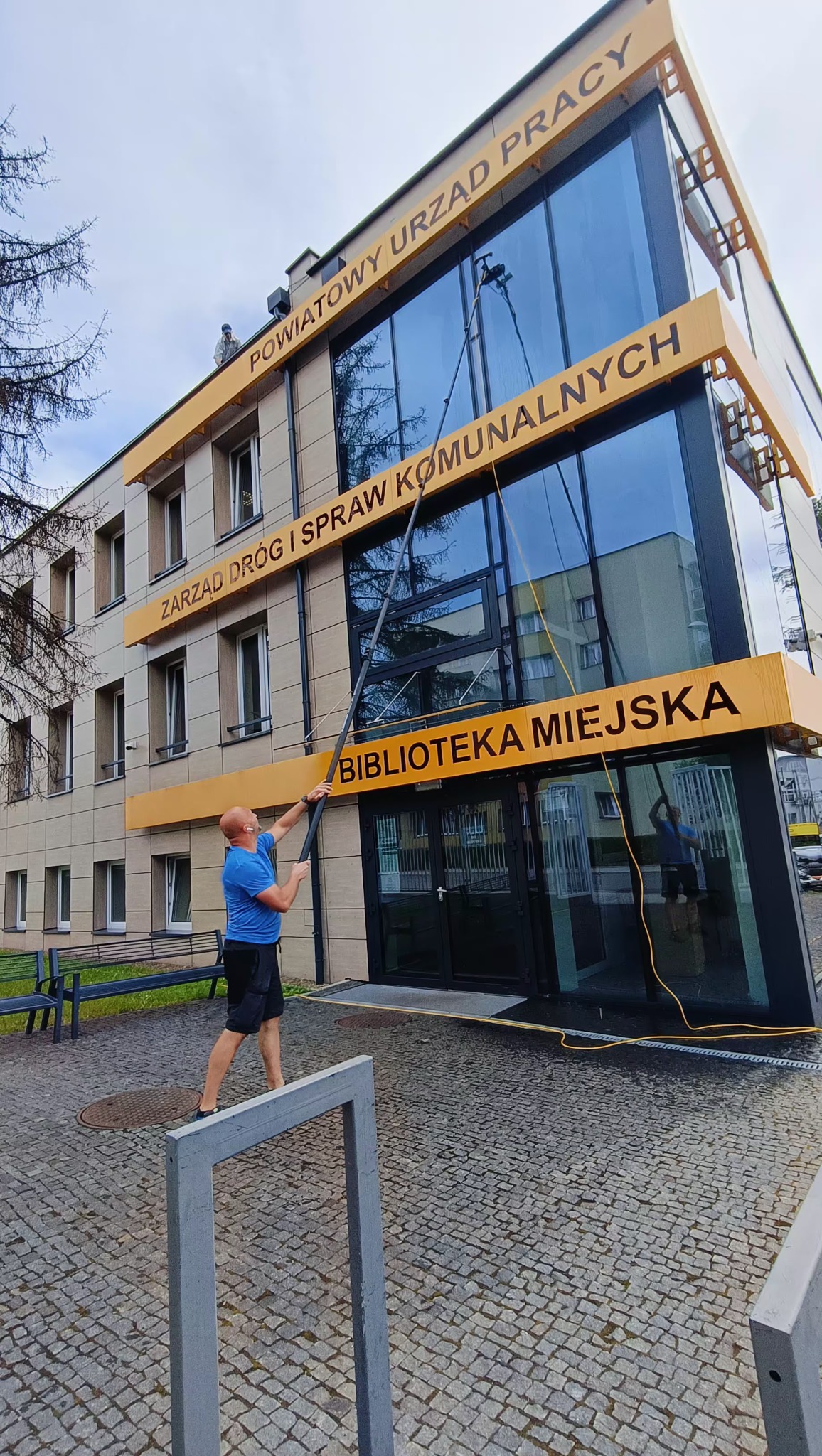 Mężczyzna w niebieskiej koszulce czyści okna Biblioteki Miejskiej za pomocą długiej lancy z dyszą, widoczny drugi pracownik na dachu budynku.