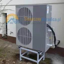 Szara jednostka zewnętrzna z dwoma wentylatorami marki OTECHNIX, zamontowana na metalowej konstrukcji przy ścianie budynku. Widoczne rury w izolacji i przewody elektryczne. Na ścianie budynku...