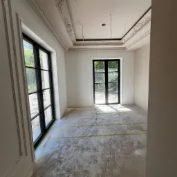 Pokój w trakcie remontu: zabezpieczona podłoga, świeżo położona gładź, dekoracyjne listwy sufitowe i okienne, czarne okna z widokiem na zieleń.