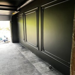 IDmal - Eleganckie, czarne ściany z dekoracyjnymi ramami w nowo wykończonym wnętrzu, widok na otwartą bramę garażową i surową posadzkę.