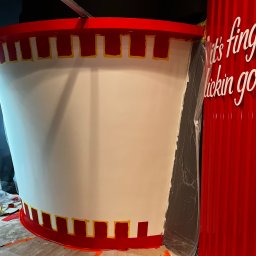 IDmal - Wnętrze lokalu w trakcie malowania dekoracji imitującej kubełek KFC, z widocznymi taśmami malarskimi i napisem reklamowym na czerwonej ścianie.
