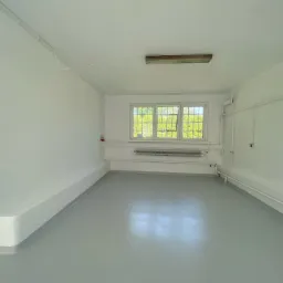 Puste, świeżo pomalowane pomieszczenie biurowe z szarym podłożem, białe ściany, okno z widokiem na zieleń, widoczne rury i grzejnik.