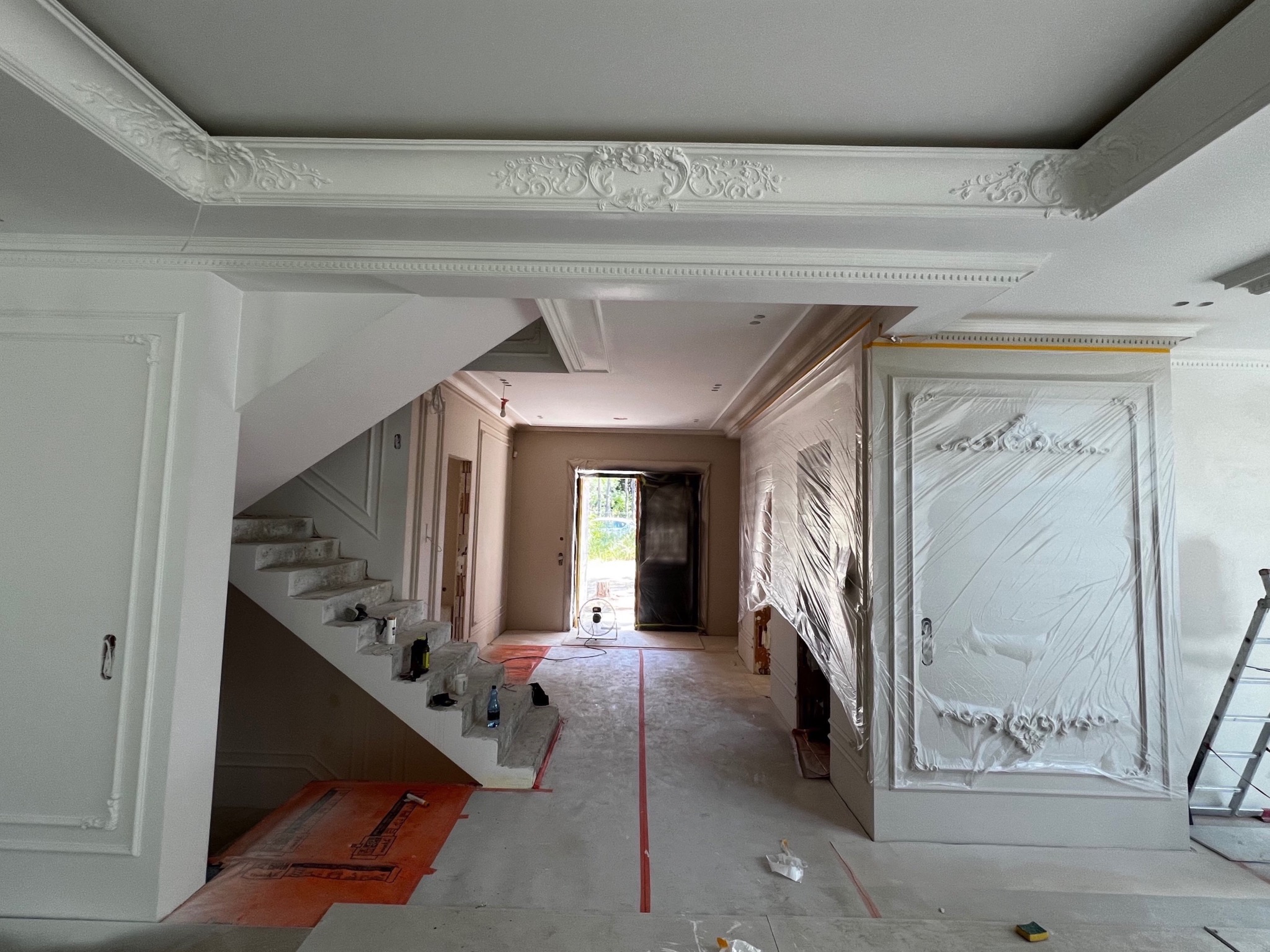 Wnętrze w trakcie remontu: białe ściany z ozdobnymi sztukateriami, schody, zabezpieczone folią meble. Widoczny etap prac wykończeniowych przed malowaniem.