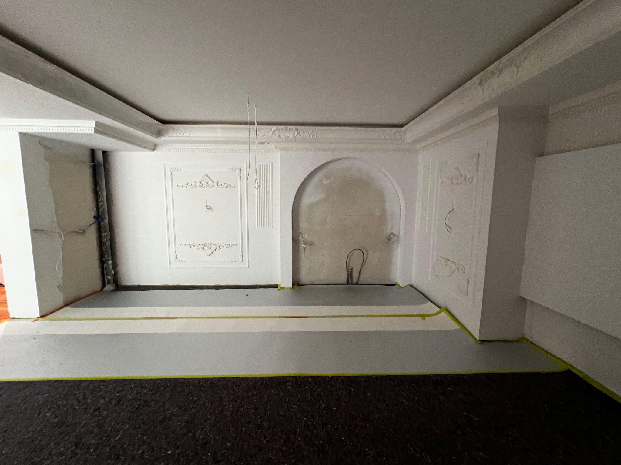 Białe ściany z dekoracyjnymi sztukateriami, przygotowane do malowania. Zabezpieczona podłoga folią i taśmą malarską. Wnęka w ścianie w trakcie szpachlowania.