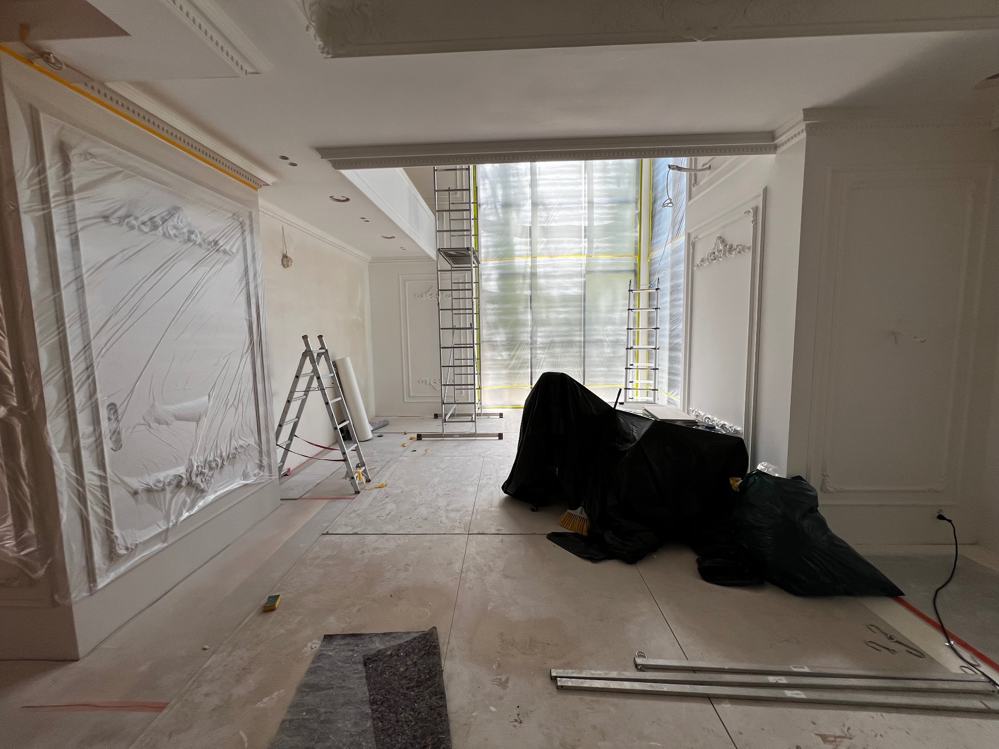 Wnętrze w trakcie prac wykończeniowych: zabezpieczone ściany z dekoracyjnymi sztukateriami, drabiny, materiały budowlane, przygotowanie do malowania.