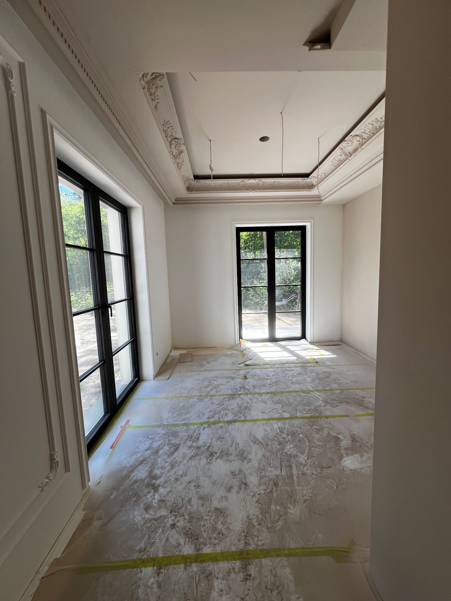 Pokój w trakcie remontu: zabezpieczona podłoga, świeżo położona gładź, dekoracyjne listwy sufitowe i okienne, czarne okna z widokiem na zieleń.