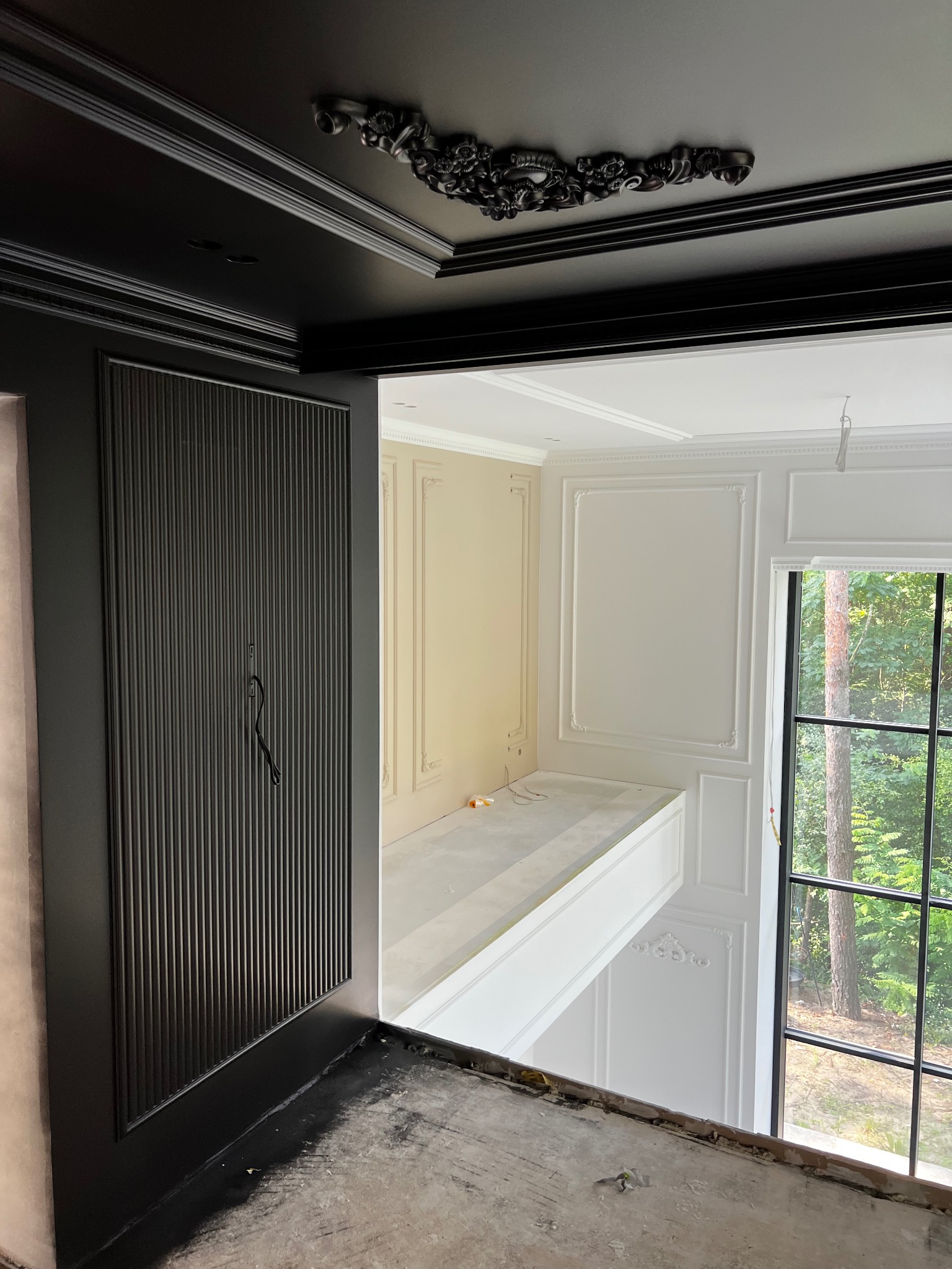 Eleganckie wnętrze w trakcie wykończenia: czarny sufit z dekoracyjną sztukaterią, panel ścienny i białe ściany z listwami. Widok na okno z zielenią w tle.