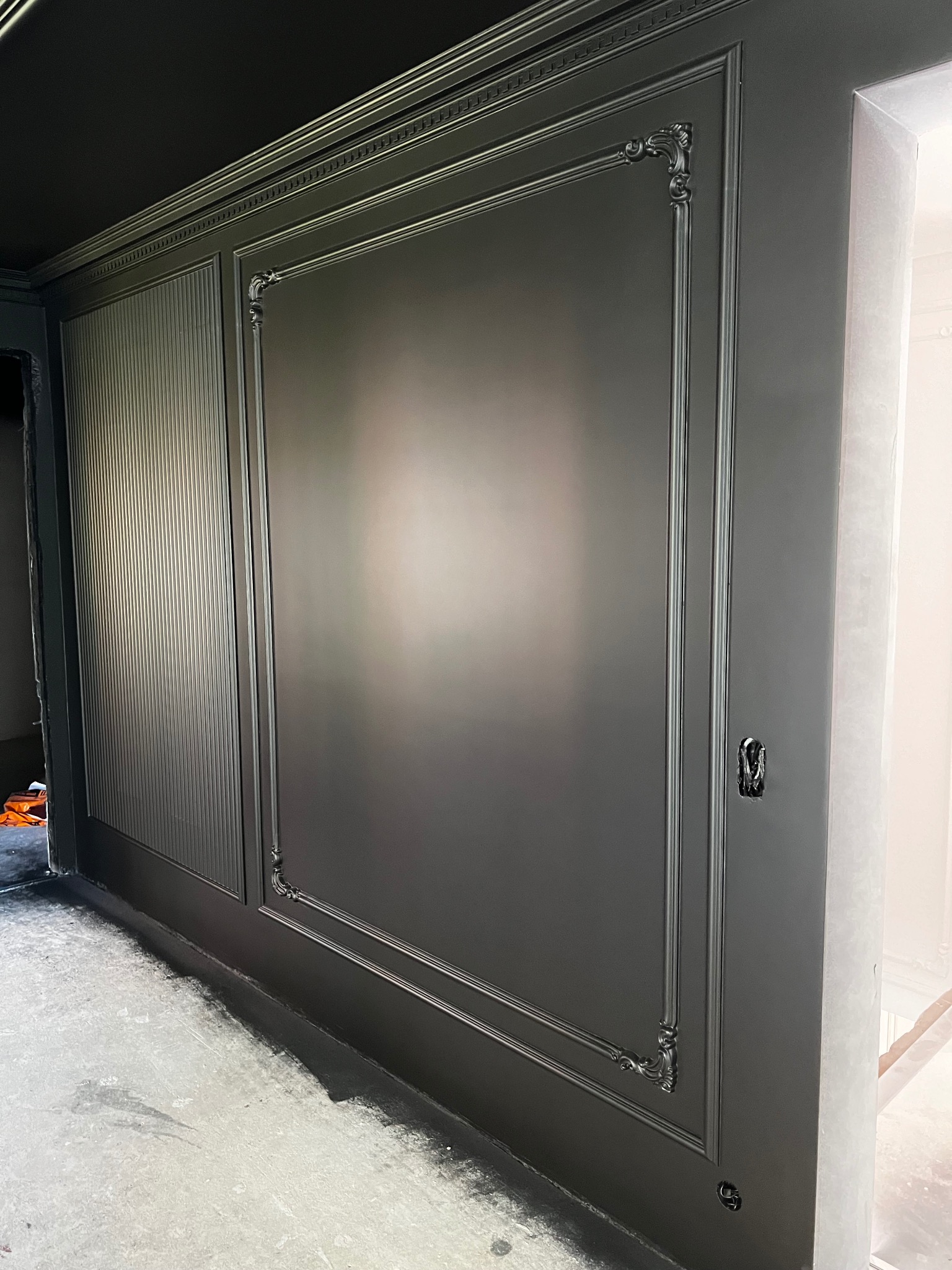 Eleganckie, ciemnoszare panele ścienne z dekoracyjnymi listwami w mieszkaniu, widoczna surowa podłoga przed wykończeniem. Detal gniazdka elektrycznego.